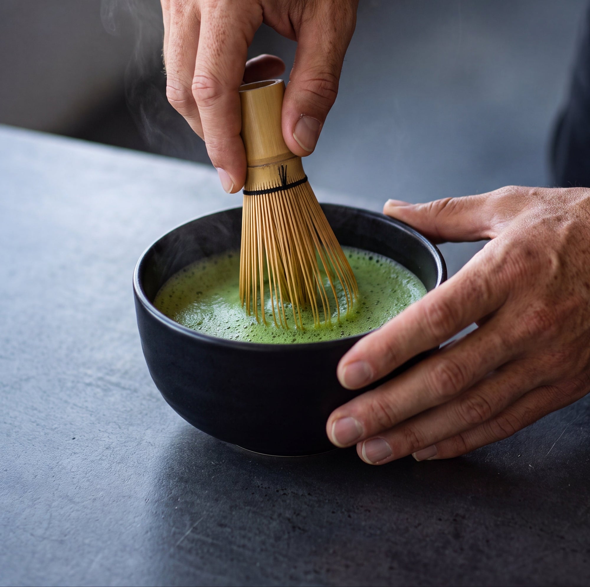 A close-up lifestyle shot of a person whisking green matcha tea in a black ceramic bowl using a bamboo chashaku. The person's hands and arms are visible, along with a bright yellow-green wristband, set against a dark gray background.